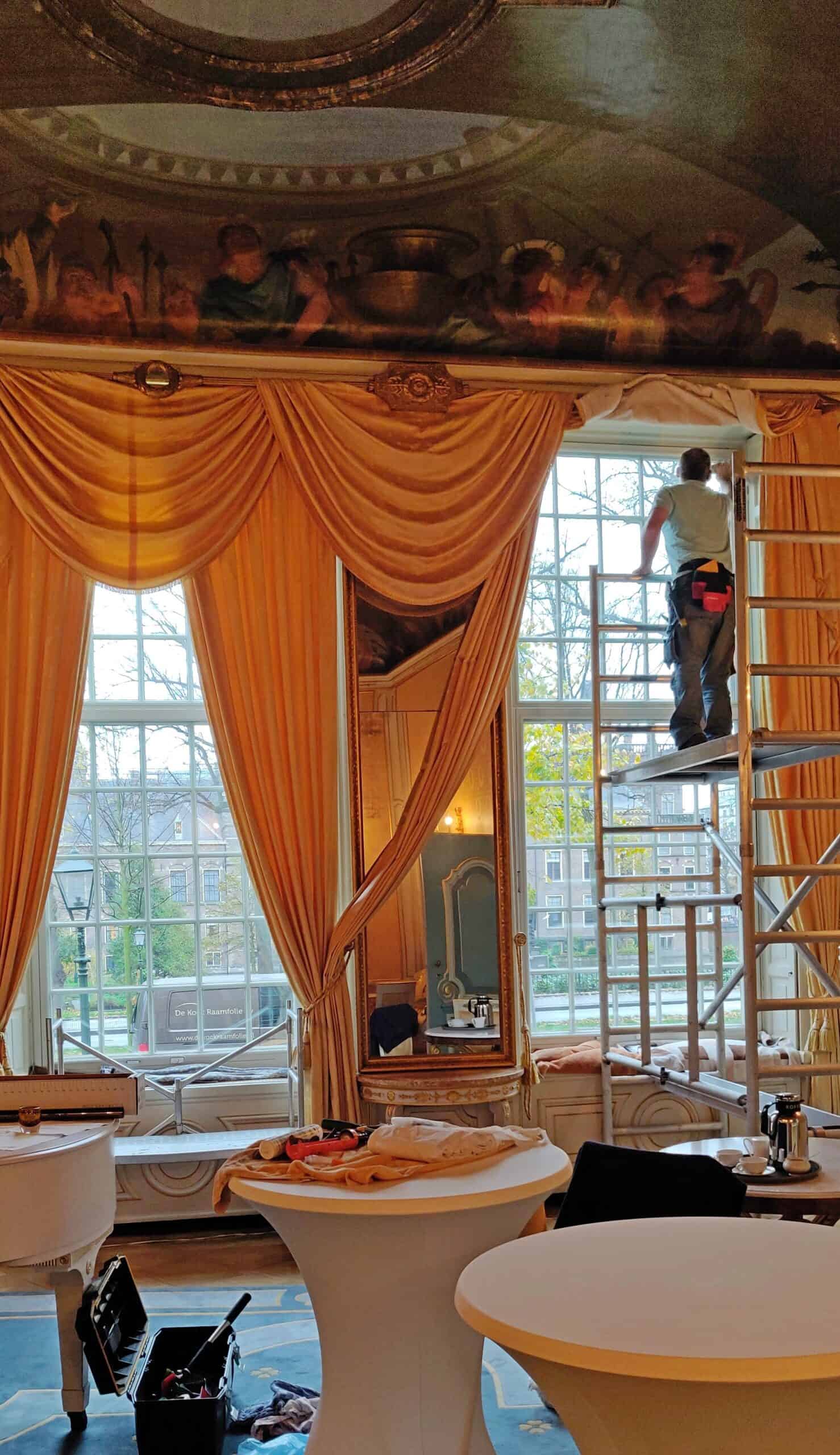 Een klassiek hoge kamer met chique gordijnen en een prachtig geschilderd plafond. Op de steiger is onze monteur bezig met het aanbrengen van zonwerende raamfolie.