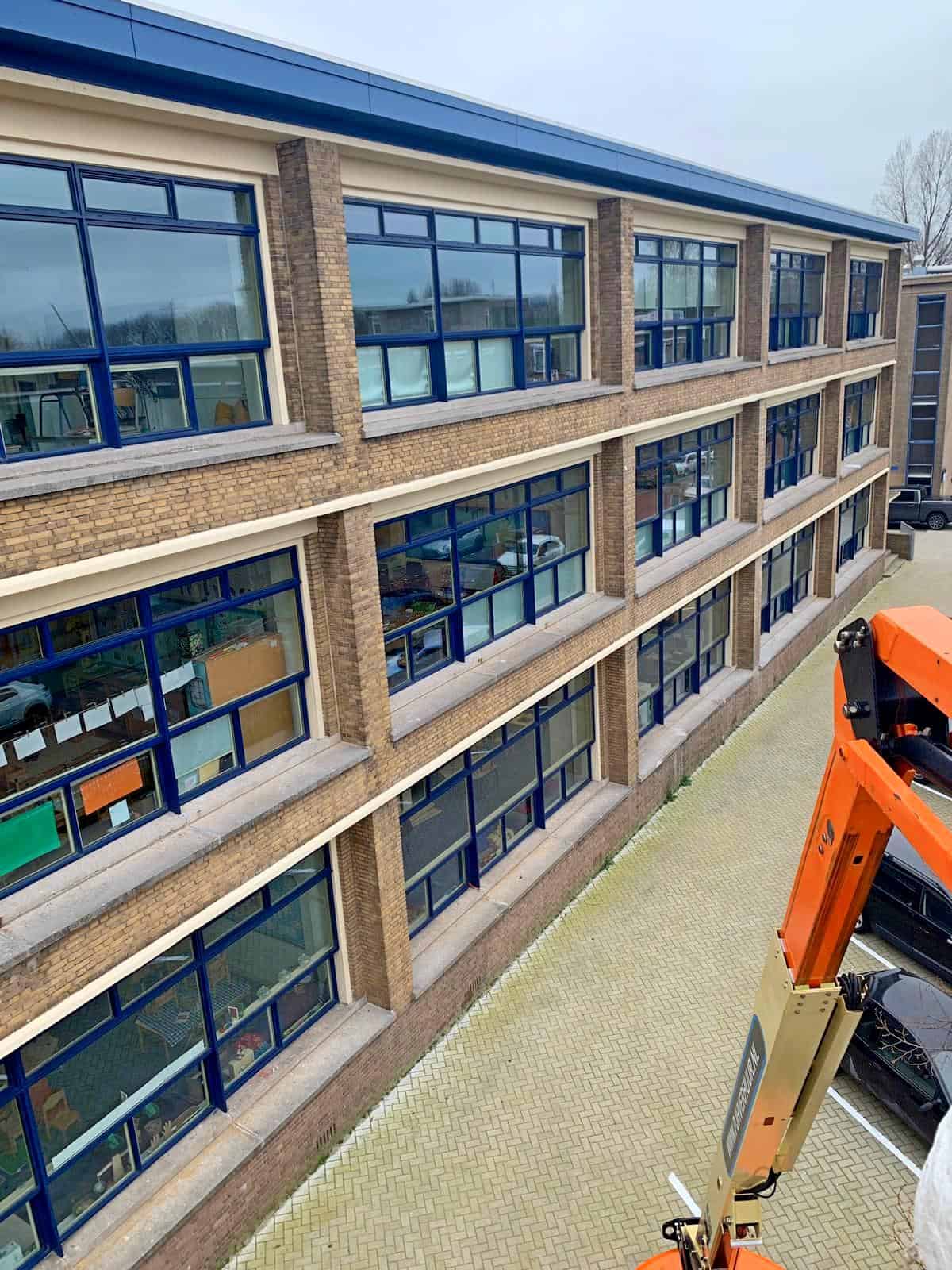 Vanuit een hoogwerker langs de gevel van de International School in Den Haag, met opvallende blauwe kozijnen. Op de voorgrond is een deel van de oranje hoogwerker zichtbaar.