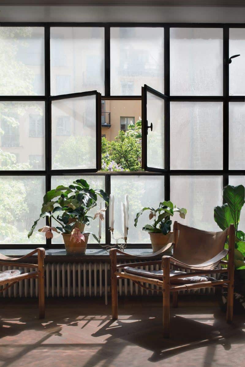 Ash Transparent 2 Een interieurfoto van drie ramen in een huis, elk voorzien van decoratief glas in de bovenste delen, met een kleurrijk patroon van gele en oranje tinten. Het onderste gedeelte van de ramen heeft matte folie, die privacy biedt terwijl het uitzicht naar buiten behouden blijft. De ramen zijn typisch voor een oud huis, met een klassieke uitstraling door de combinatie van het glas-in-lood boven en het moderne matte folie eronder. In de achtergrond zijn andere huizen zichtbaar.